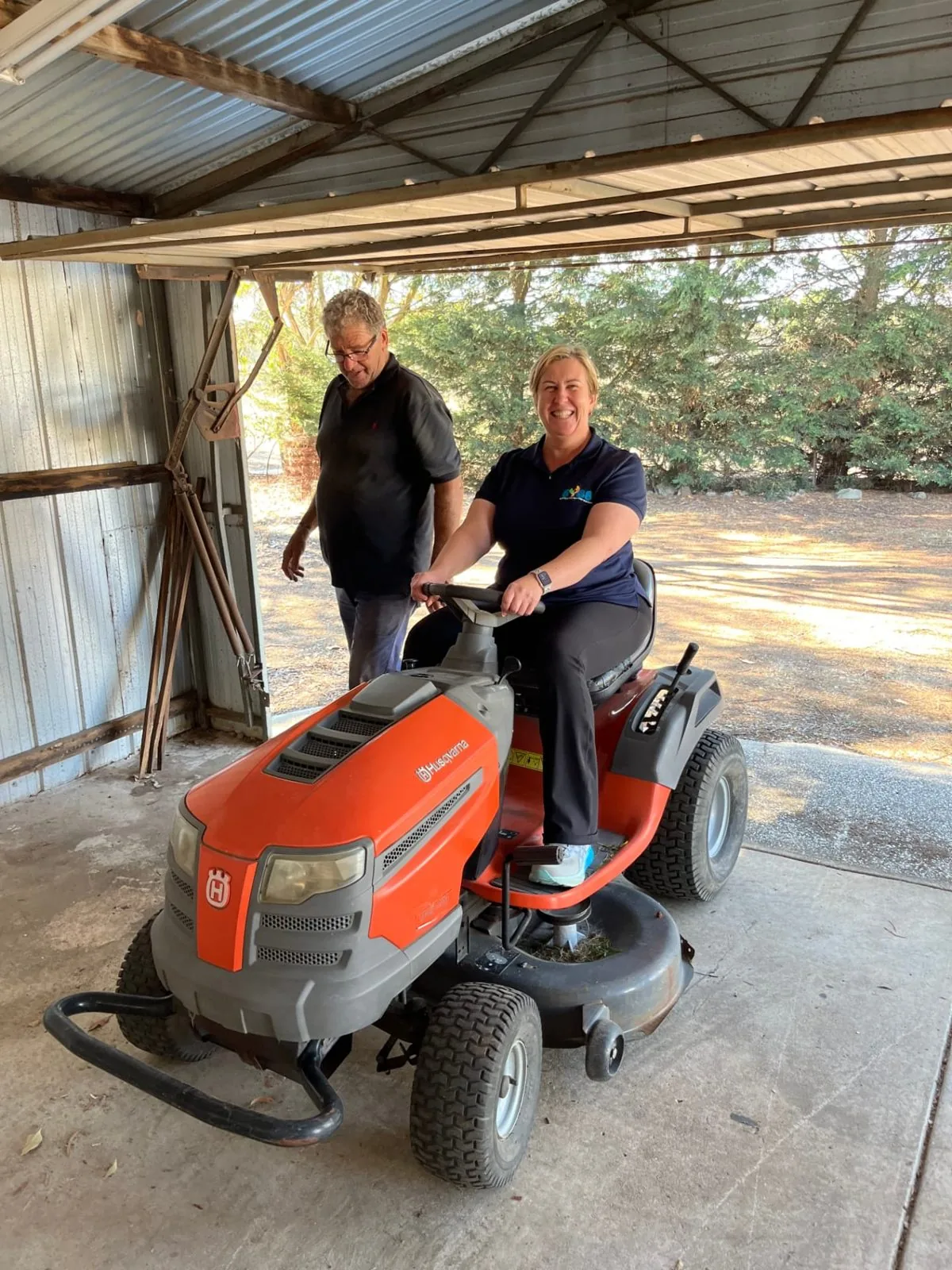 Support worker on a ride-on mower sharing a laugh with a participant in the shed